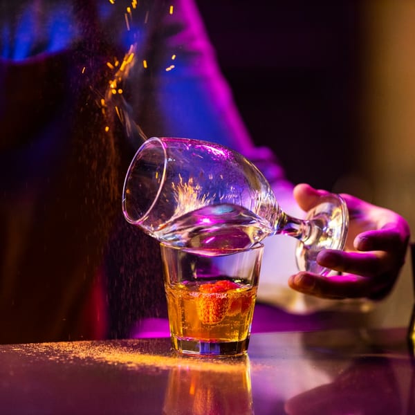 A bartender ignites a drink with sparks flying, illuminated by vibrant purple and blue lights.