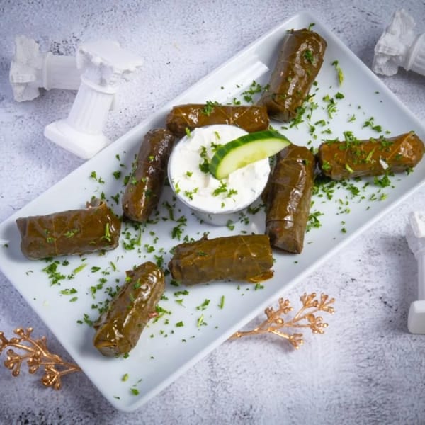 A white rectangular plate of dolmades, garnished with parsley and a side of tzatziki with cucumber.
