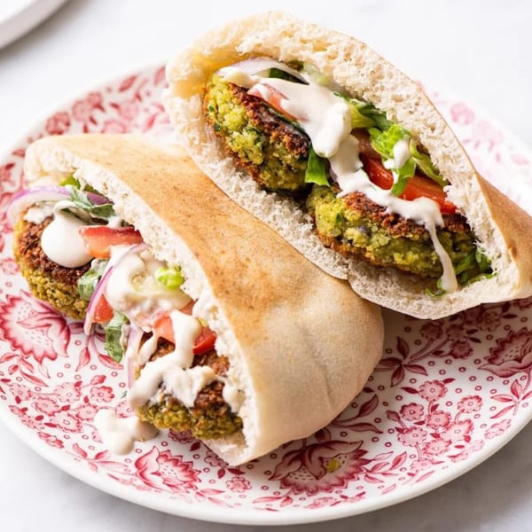 Two falafel pita sandwiches with lettuce, tomatoes, red onions, and tahini sauce on a floral plate.