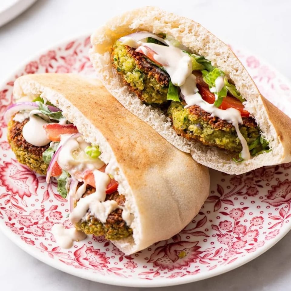 Two falafel pita sandwiches with lettuce, tomatoes, red onions, and tahini sauce on a floral plate.