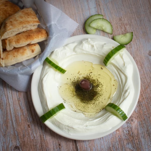 Labneh dip with olive oil, za'atar, an olive, cucumber slices, and pita bread on a wooden table.