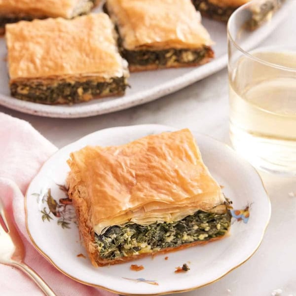 A square of golden-brown spinach and feta phyllo pastry on a decorative white plate with a drink.