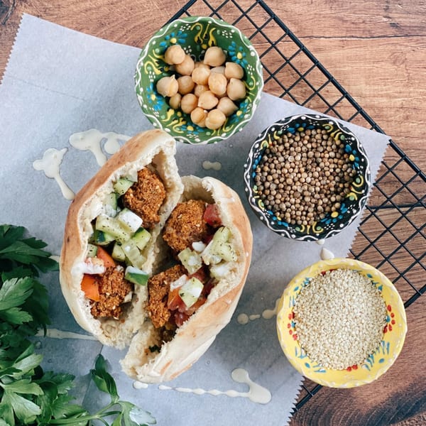 Halal falafel pita with cucumber, tomato, and tahini, alongside bowls of chickpeas, coriander, and sesame seeds.