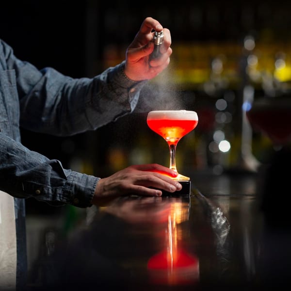 Bartender spraying a mist over a glowing red cocktail on a dark bar counter.