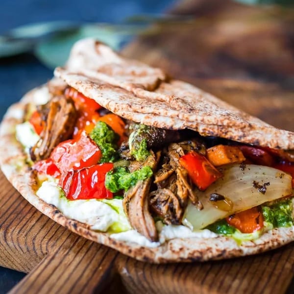 Halal Mediterranean pita wrap with meat, roasted vegetables, white sauce, and green pesto on a wooden board.