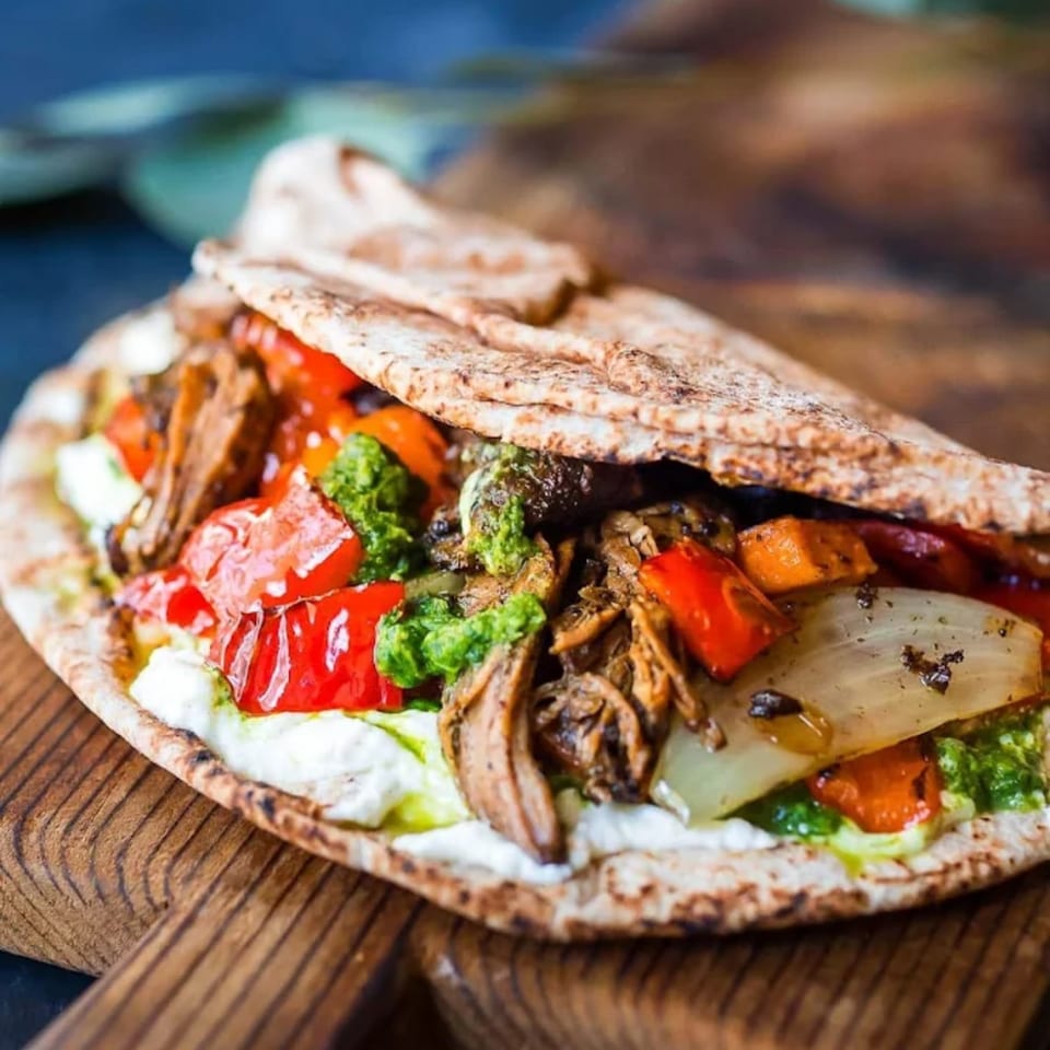 Halal Mediterranean pita wrap with meat, roasted vegetables, white sauce, and green pesto on a wooden board.